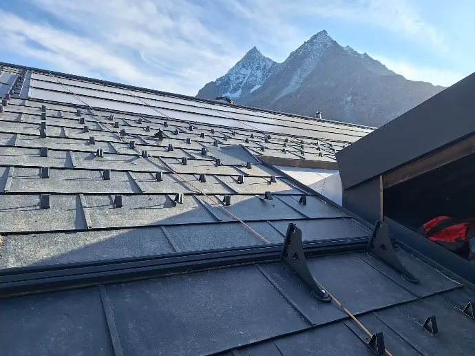 Installation d'une toiture en ardoises synthétiques ou bac acier avec système pare-neige renforcé en haute montagne. Vue sur les crochets de retenue de neige et le profil à joint debout anthracite sur un versant à forte pente face aux sommets enneigés.