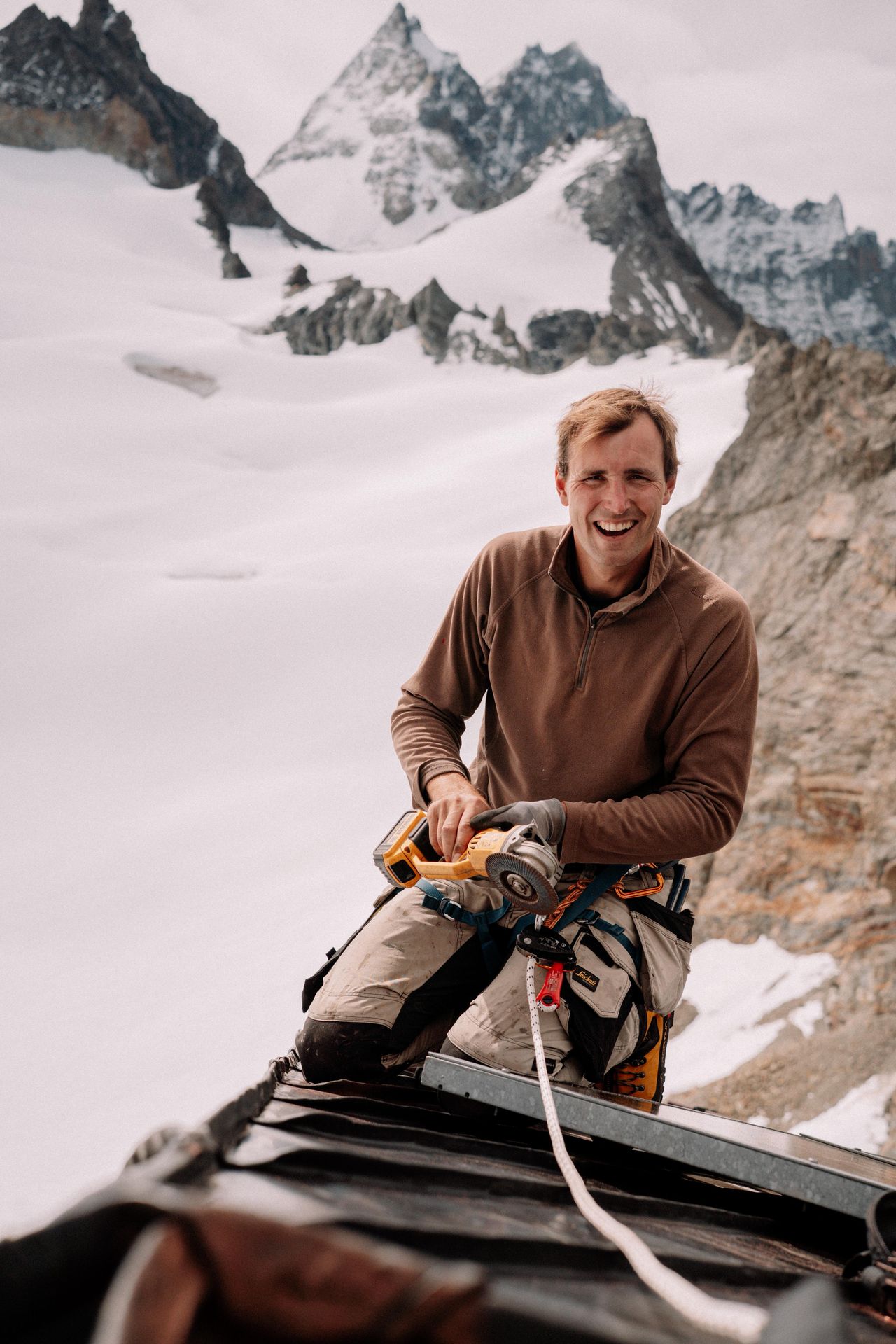 Artisan couvreur cordiste souriant en intervention de haute montagne. Utilisation d'une disqueuse portative pour la découpe de précision d'une toiture métallique en altitude. Équipement de sécurité complet et harnais pour travaux en accès difficile face à un sommet enneigé.