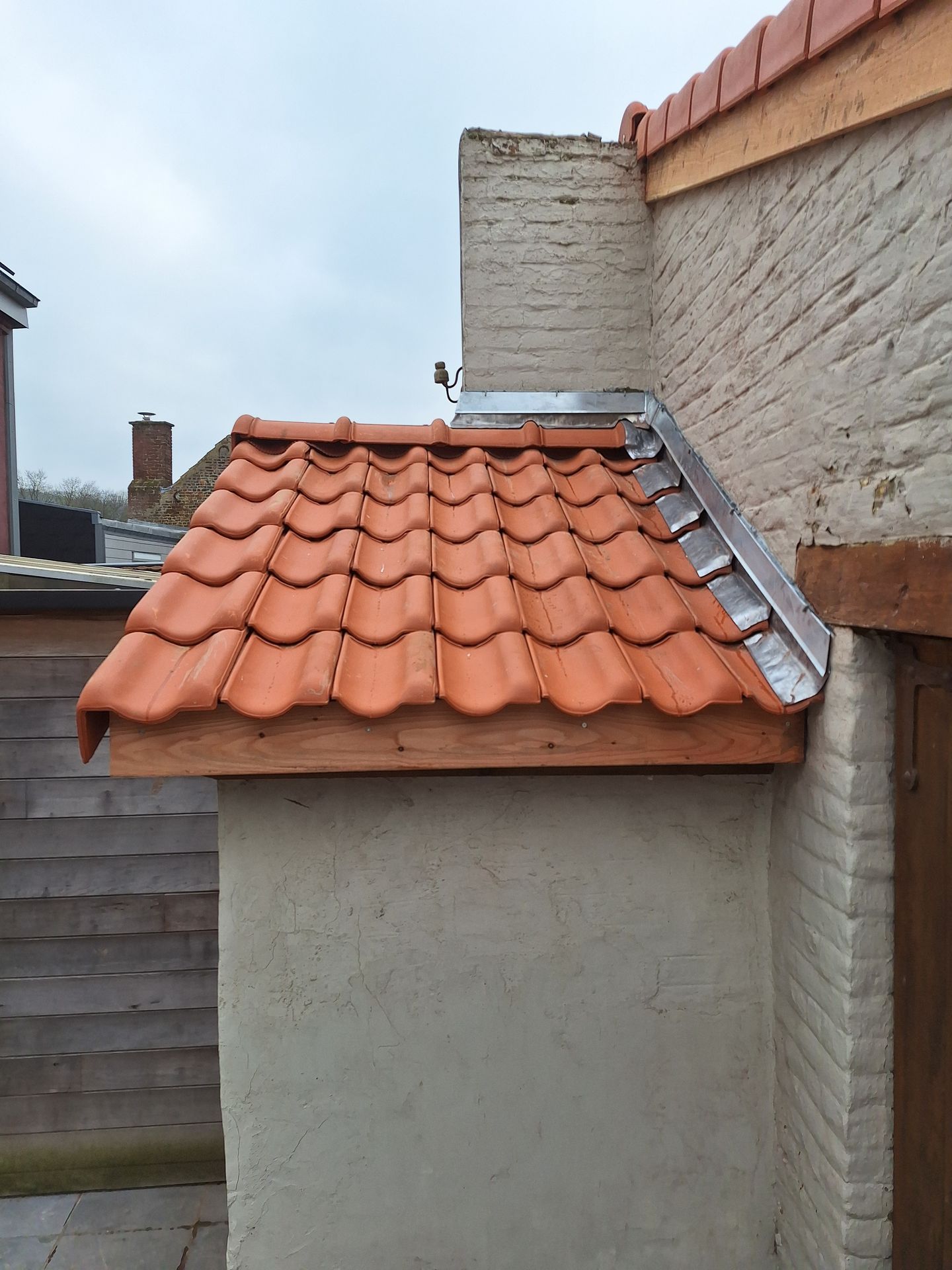 Étanchéité murale de toiture en tuiles terre cuite avec pose de solins en plomb et porte-solins métalliques. Finition de rive contre mur sur une petite toiture en appentis avec structure bois apparente et faîtage maçonné.