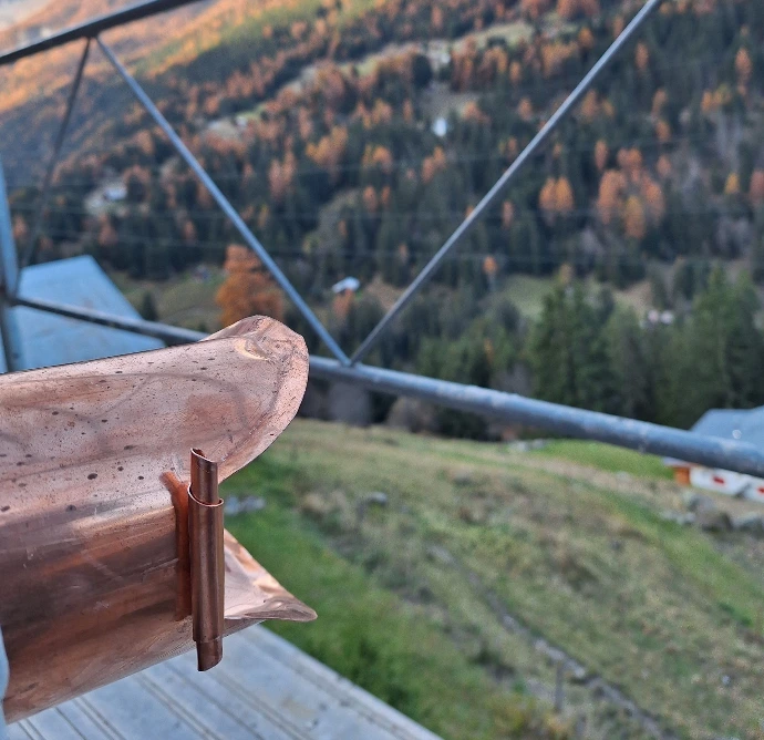 Détail technique d'un élément de zinguerie en cuivre réalisé sur-mesure pour une toiture de montagne. Vue sur la finition artisanale d'un chéneau ou d'un habillage métallique avec paysage alpin en arrière-plan. Travail de précision sur métaux nobles pour une longévité maximale.