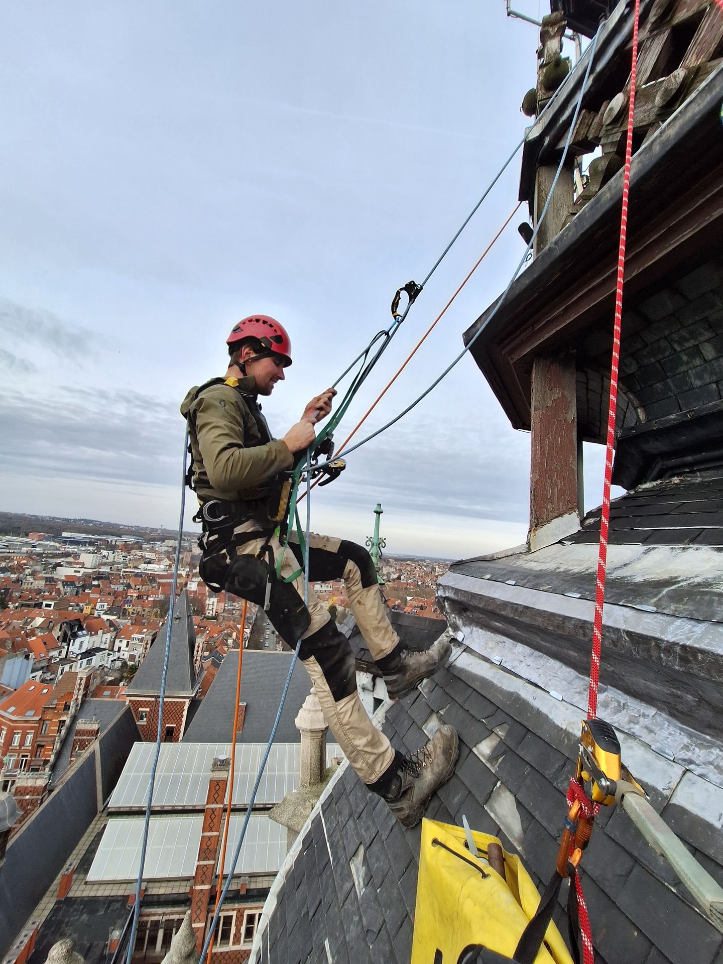 Cordiste professionnel effectuant des travaux de zinguerie sur une toiture en ardoise à forte pente. Sécurisé par des cordes, descendeur et mousquetons sur dispositif d'ancrage, au-dessus d'une ville.
