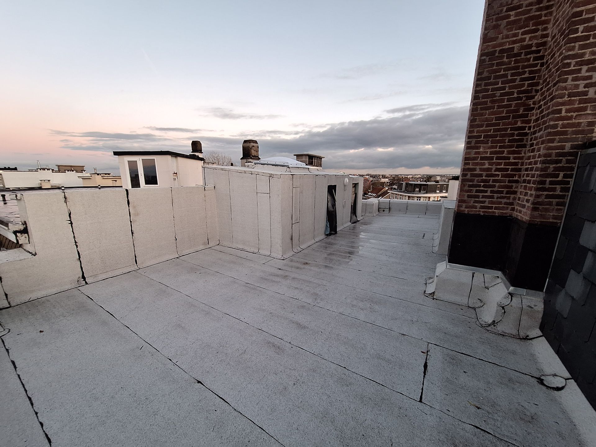 Étanchéité de toiture terrasse avec membrane bitumineuse grise sur un toit plat urbain. On aperçoit les relevés d'étanchéité sur les acrotères, une souche de cheminée en briques avec solins, et un lanterneau d'éclairage zénithal au second plan.