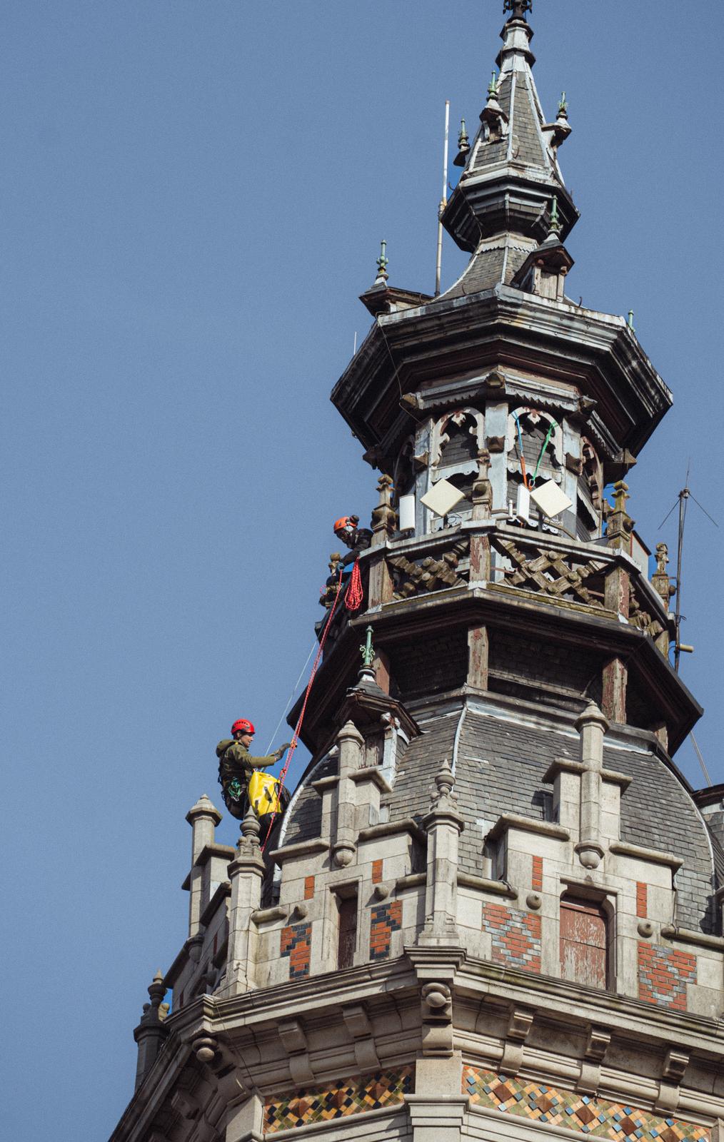 Intervention de cordistes experts sur un clocher historique ou tour ornementale. Travaux de restauration de toiture en ardoises naturelles et zinguerie d'art. Accès difficile par cordes sur une structure de prestige avec détails architecturaux anciens et flèche de clocher.