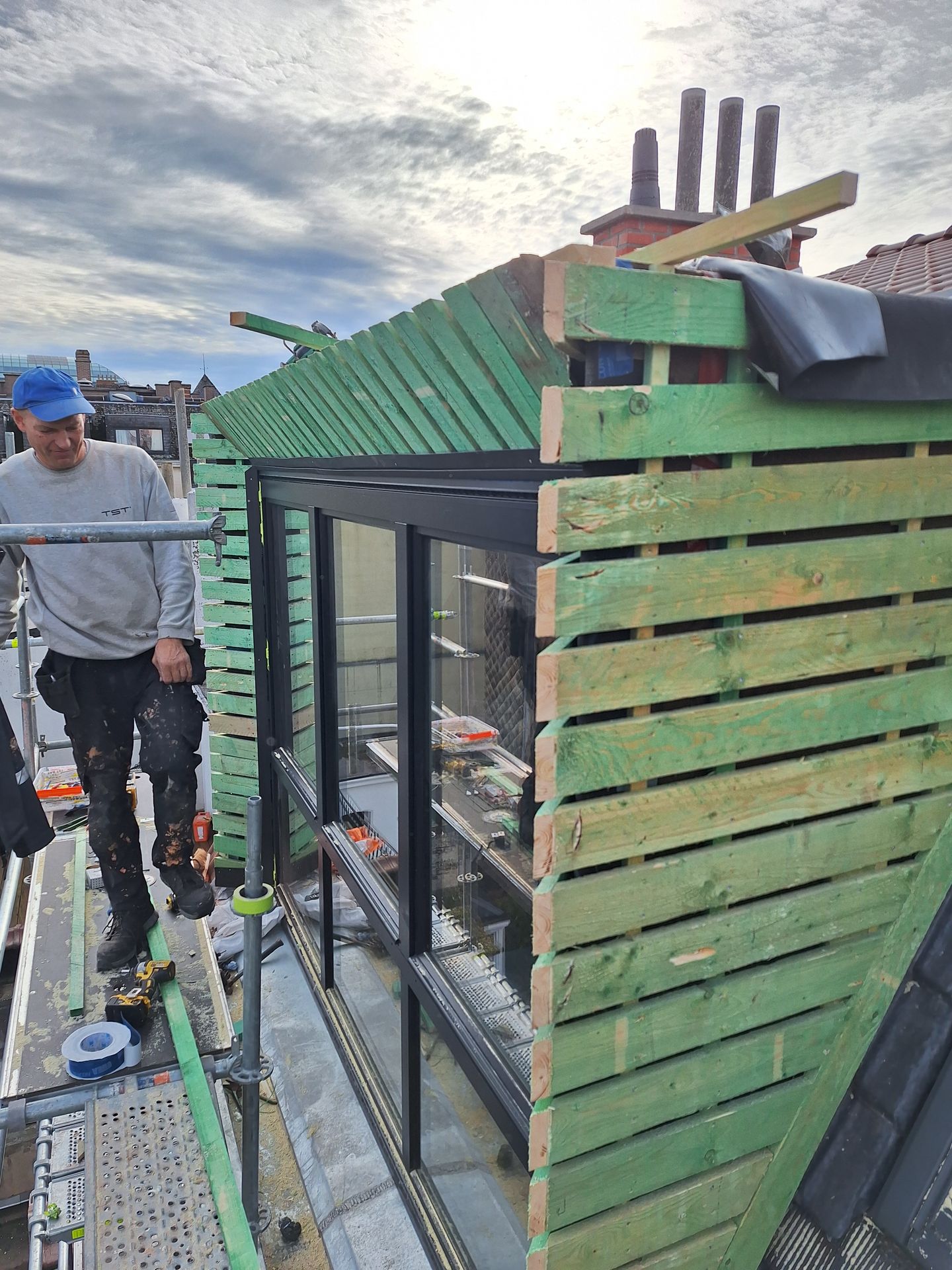 Chantier de création d'une lucarne rampante : pose du contre-lattage en bois traité sur ossature, intégration d'un châssis vitré fixe et ouvrant. Travaux sur échafaudage de pied avec vue sur les conduits de cheminée et l'isolation périphérique avant la pose du bardage en zinc.