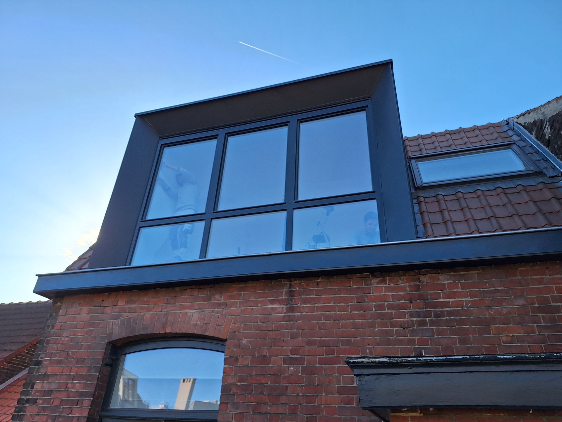 Installation d'une lucarne rampante moderne en zinc avec de grandes baies vitrées sur une toiture en tuiles terre cuite. Finitions de zinguerie sur-mesure pour l'étanchéité de la structure et intégration d'une fenêtre de toit de type Velux sur le versant adjacent d'une maison en briques