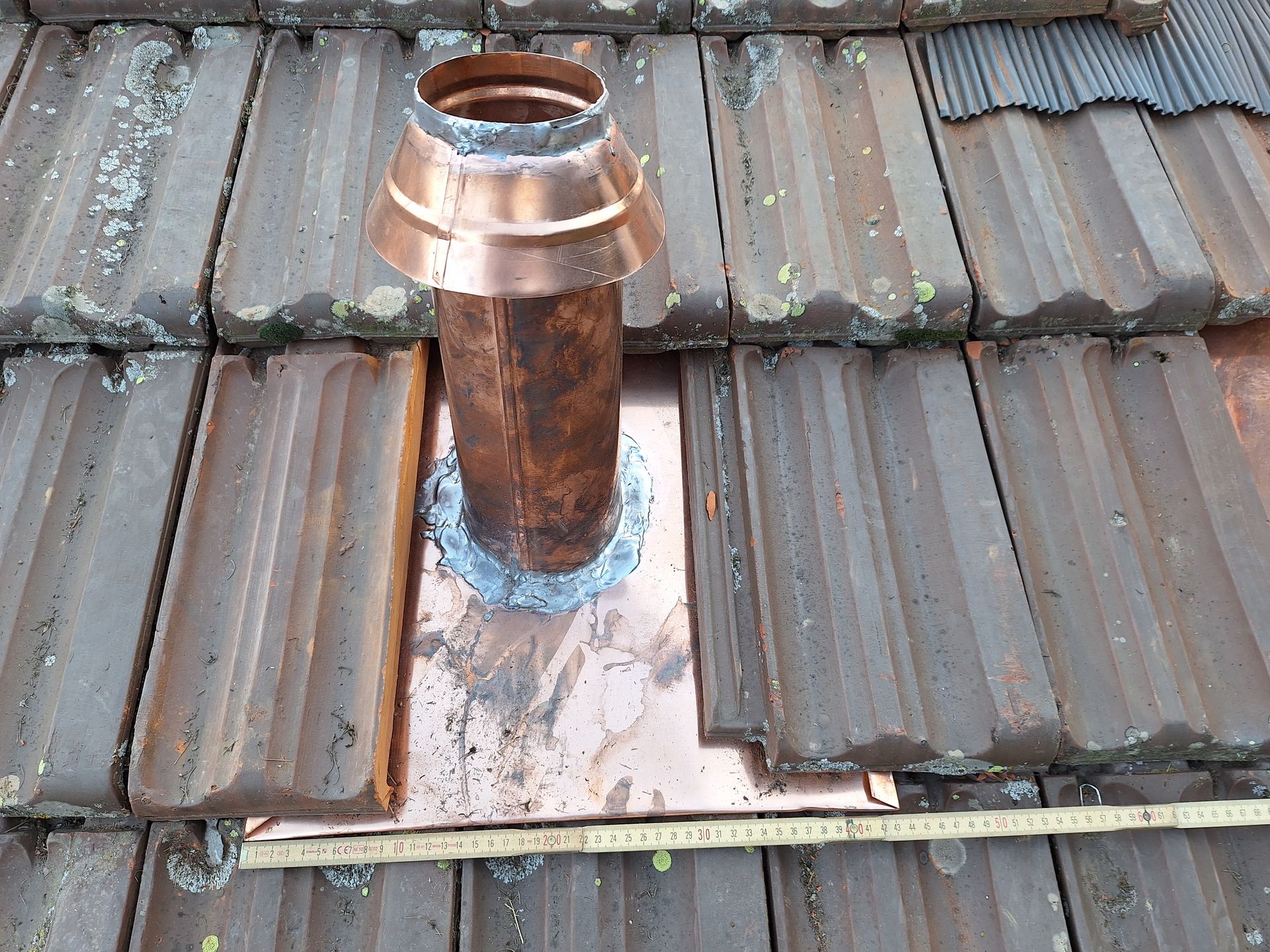 Raccord d'étanchéité de sortie de cheminée en cuivre sur toiture en tuiles terre cuite. Pose d'une collerette de finition et d'une plaque de cuivre sur-mesure avec soudures à l'étain. Détail technique d'un abergement de toiture pour une évacuation étanche.