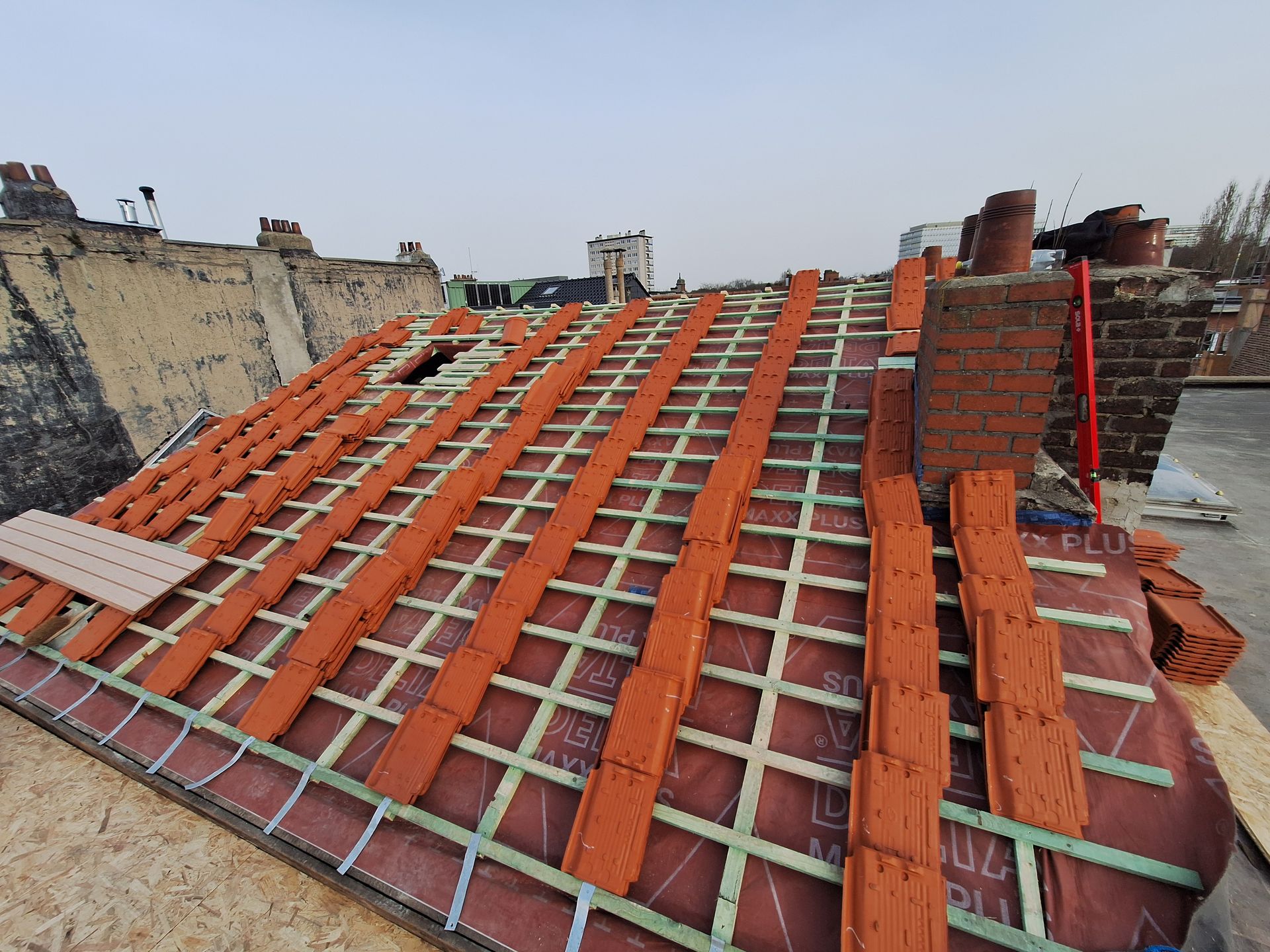 Chantier de rénovation de toiture : pose de tuiles terre cuite sur lattage et contre-lattage. Vue sur l'écran sous-toiture HPV (Haute Perméabilité à la Vapeur), les crochets de sécurité et l'abergement de cheminée en briques en cours de réalisation sur un versant à forte pente.