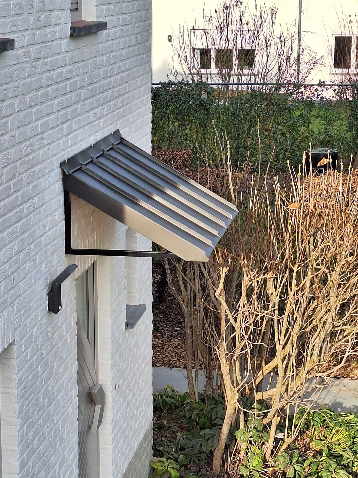 Auvent de porte d'entrée moderne avec couverture en zinc à joint debout anthracite sur structure métallique noire. Pose en façade sur mur de briques blanches avec étanchéité par solin métallique et raccord de zinguerie sur-mesure pour protection de seuil.