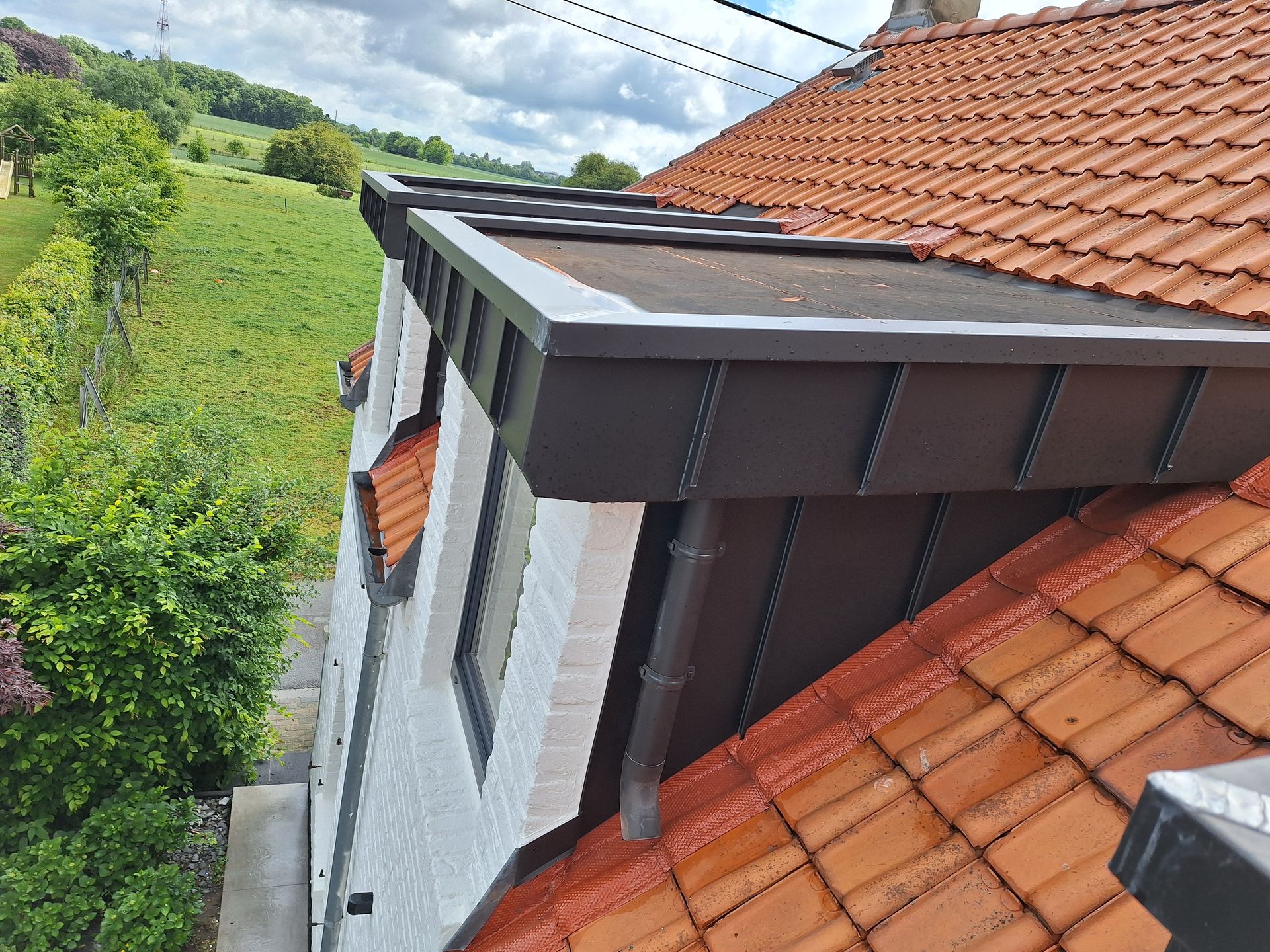 Habillage de lucarne rampante en zinc joint debout anthracite avec raccord d'étanchéité sur toiture en tuiles terre cuite. Vue sur la rive de toit, le chéneau de récupération des eaux pluviales et la descente de gouttière intégrée. Travail de zinguerie de précision pour extension de toiture.
